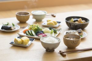 Repas japonais posé sur une table en bois, composé de bol de riz et soupe miso, assiette de pickles japonais posé au centre avec des omelettes japonaises posées dans des assiettes, et des baguettes qui reposent sur des porte-baguettes devant les bols de riz