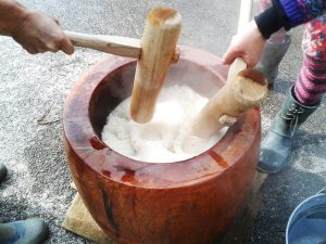 Deux personnes en train de pétrir du riz gluant, ou de la farine de riz gluante à l'aide de pilons en bois dans un grand contenant ressemblant à une cuve en bois marron. Cette cuve est posée sur un sol gris. 