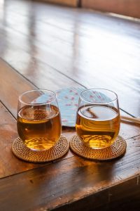 Deux verres contenant du thé froid japonais, de l'hôjicha, qui donne une couleur orangée à la boisson. Les verres sont posés sur une table en bois de couleur marron. Un éventail bleu à motif rose se trouve en arrière plan, posé sur la table également. 