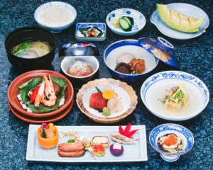 Nombreux bols et assiettes disposés sur une table bleue avec différents mets japonais colorés, comprenant des légumes, du poisson, de la viande et des fruits.