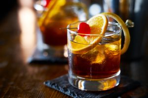 Boisson alcoolisée comme du whisky avec des glaçons, une tranche de citron sur le dessus, cerise confite sur dessous de verre en ardoise posé sur une table en bois