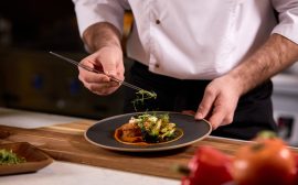 Focus sur les mains d'un chef qui dresse un plat de viande et légumes dans une assiette noire posée sur une planche en bois