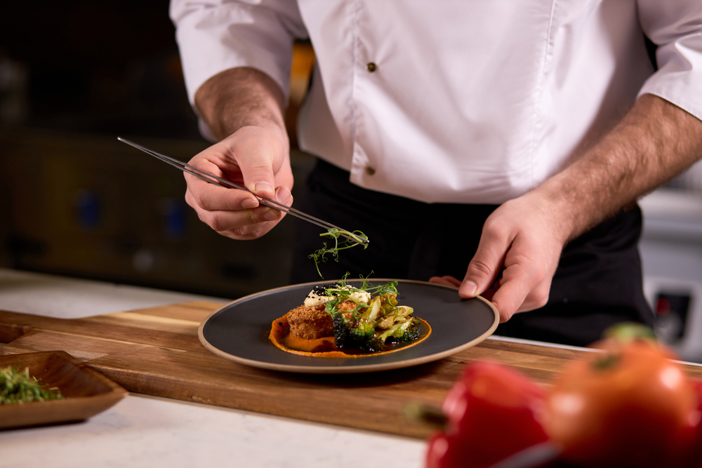 Focus sur les mains d'un chef qui dresse un plat de viande et légumes dans une assiette noire posée sur une planche en bois