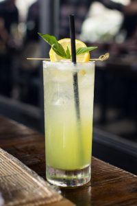 Plan fixe sur un long verre avec boisson au yuzu, une paille noire, une tranche de citron au-dessus des glaçons, posé sur un comptoir de bar en bois