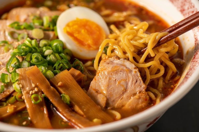 Focus sur un bol de ramen au miso, avec les baguettes qui tiennent les nouilles, un oeuf, de la ciboule et une tranche de viande