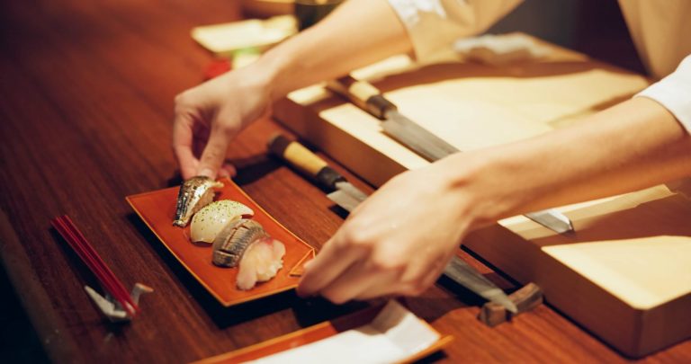 Un maître sushi qui pose un plateau de quatre sushis sur un comptoir en bois, devant lequel sont posées des baguettes